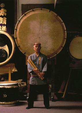 Man stands in traditional dress in front of a large drum head